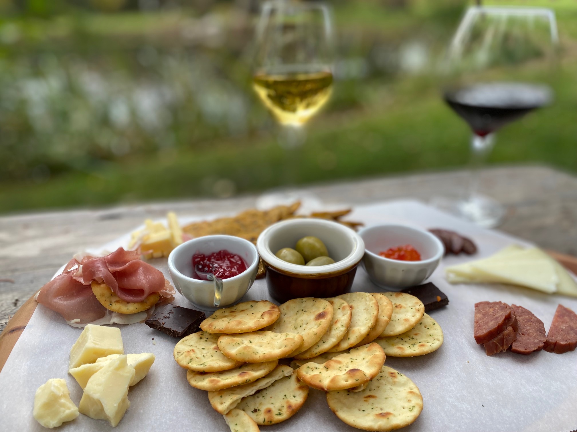 Charcuterie board of cured meats, cheese, crackers and olives with red wine and white wine on a tranquil lakeside table. Rural Route Tour Company tour.